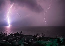 Lightening strikes water in background. Aircraft carrier with jets sitting on it in foreground.