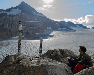 Student sitting alongside large rocks with scientific equipment set up around him, large glacier and mountains in the background.