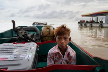 A young boy sits in a boat on top of brackish waters, another boat in the background.