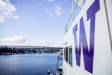 The side of a large boat with a purple UW "W" painted on it, water and bridge in background.