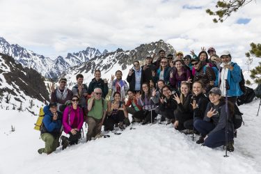 A group of about 30 students in hiking gear pose with their professor in the snow at the top of a Cascade mountain peak