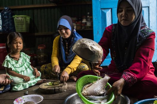 Two women in hijabs and a toddler sit cross-legged on the floor and prepare dinner. One washes fish