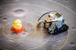 A Lego Mindstorms robot, with a plastic astronaut strapped to the front, approaches the lunar lander.
