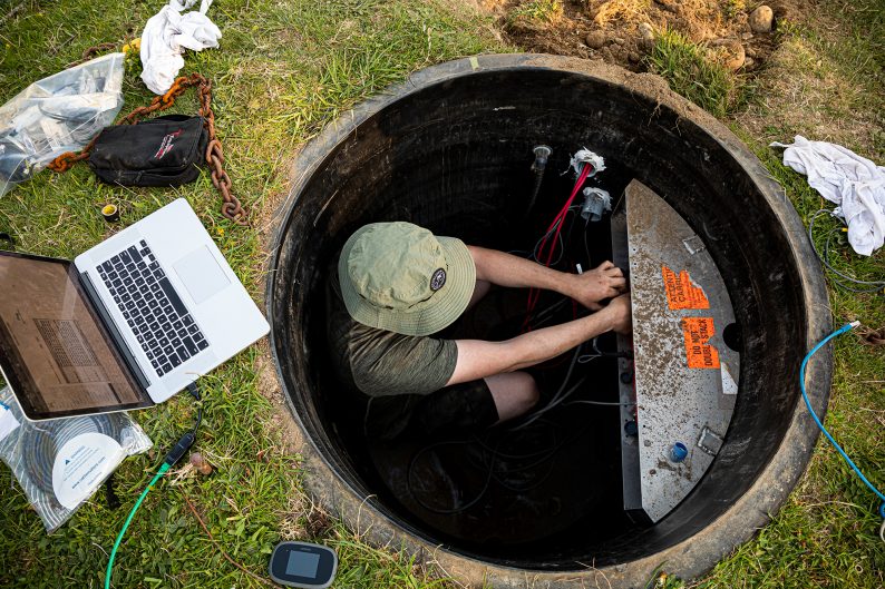 A man adjusts equipment down in a 4 foot wide hole in the ground. A laptop sits on the ground nearby.