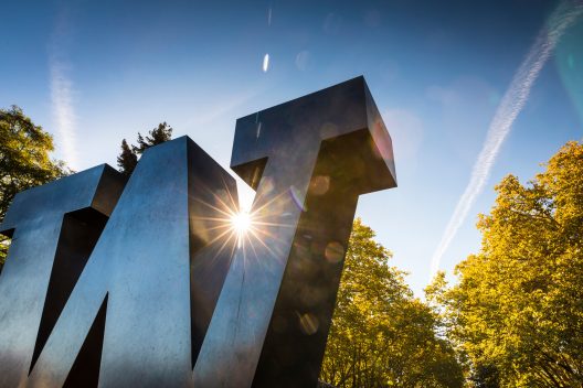A view of the large metal W on the Seattle Campus with the sun peeking out behind.